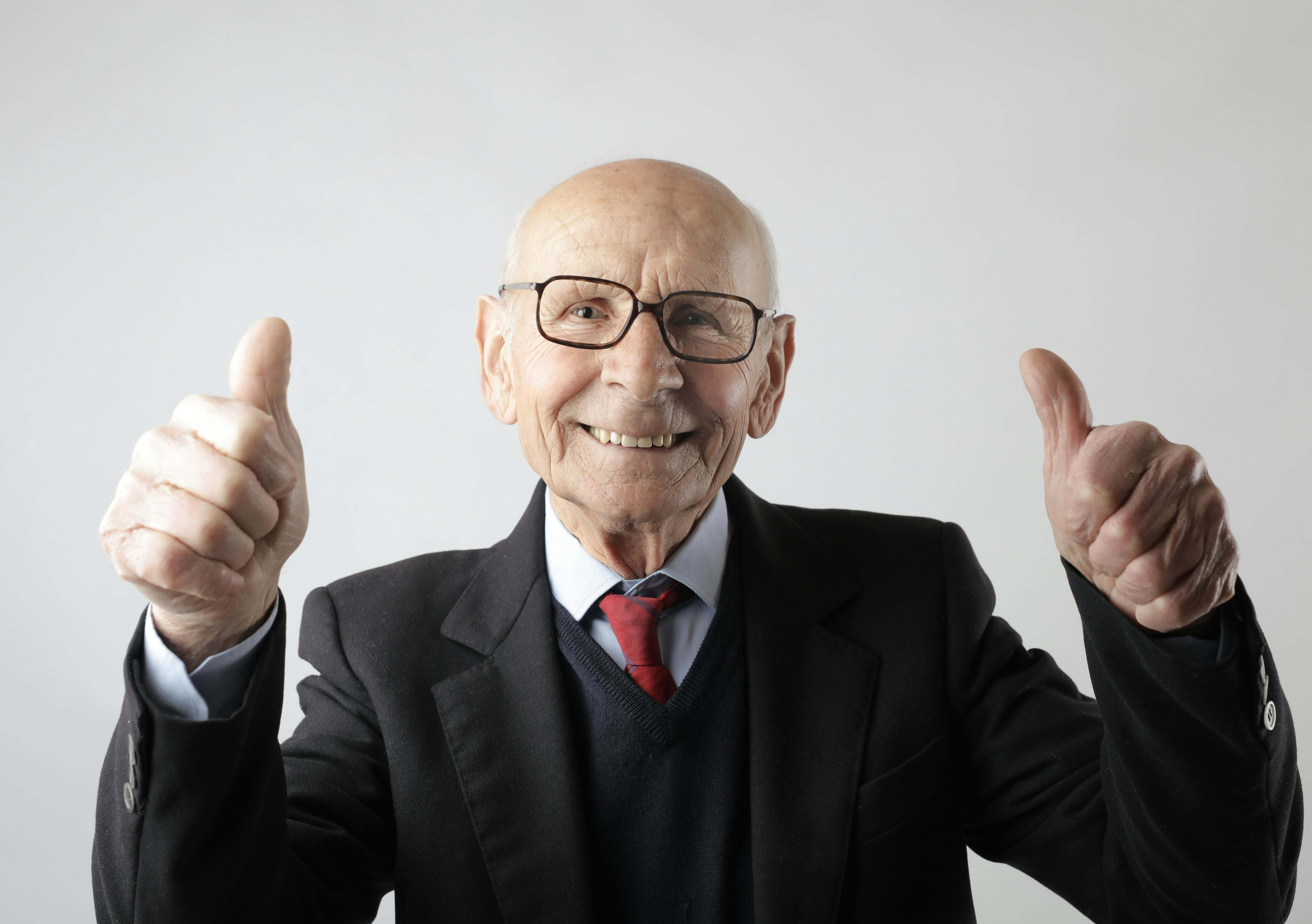 Optimistic senior man in elegant formal black suit and eyeglasses standing and showing thumbs up while smiling and looking at camera