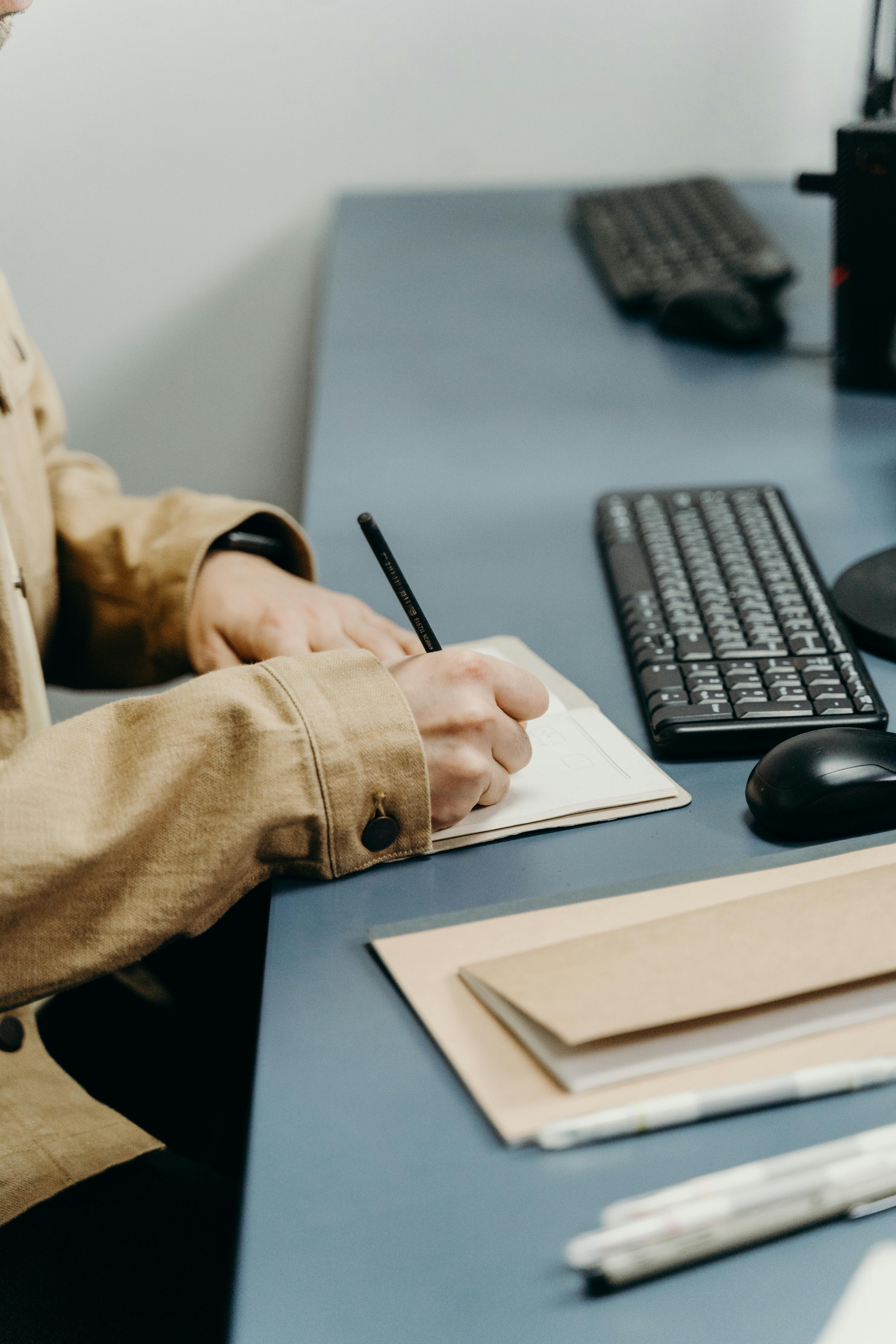 Close-up of hands writing in a notebook on an office desk with keyboard.
